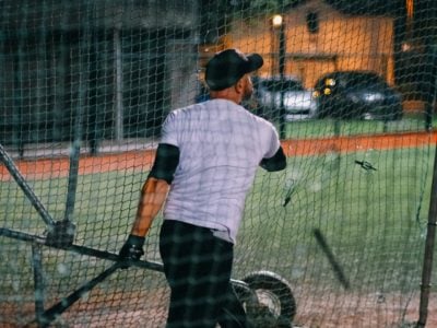Man in a batting cage wearing white t-shirt and black hat and trousers